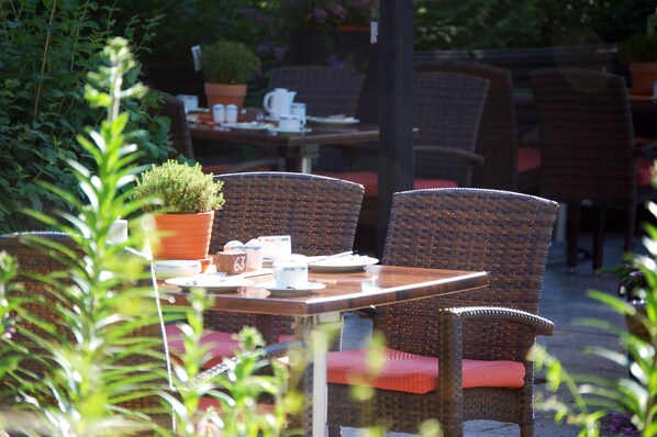 Un área de comedor al aire libre con sillas de mimbre y una mesa preparada para dos personas, rodeada de plantas en macetas y un muro de ladrillo.