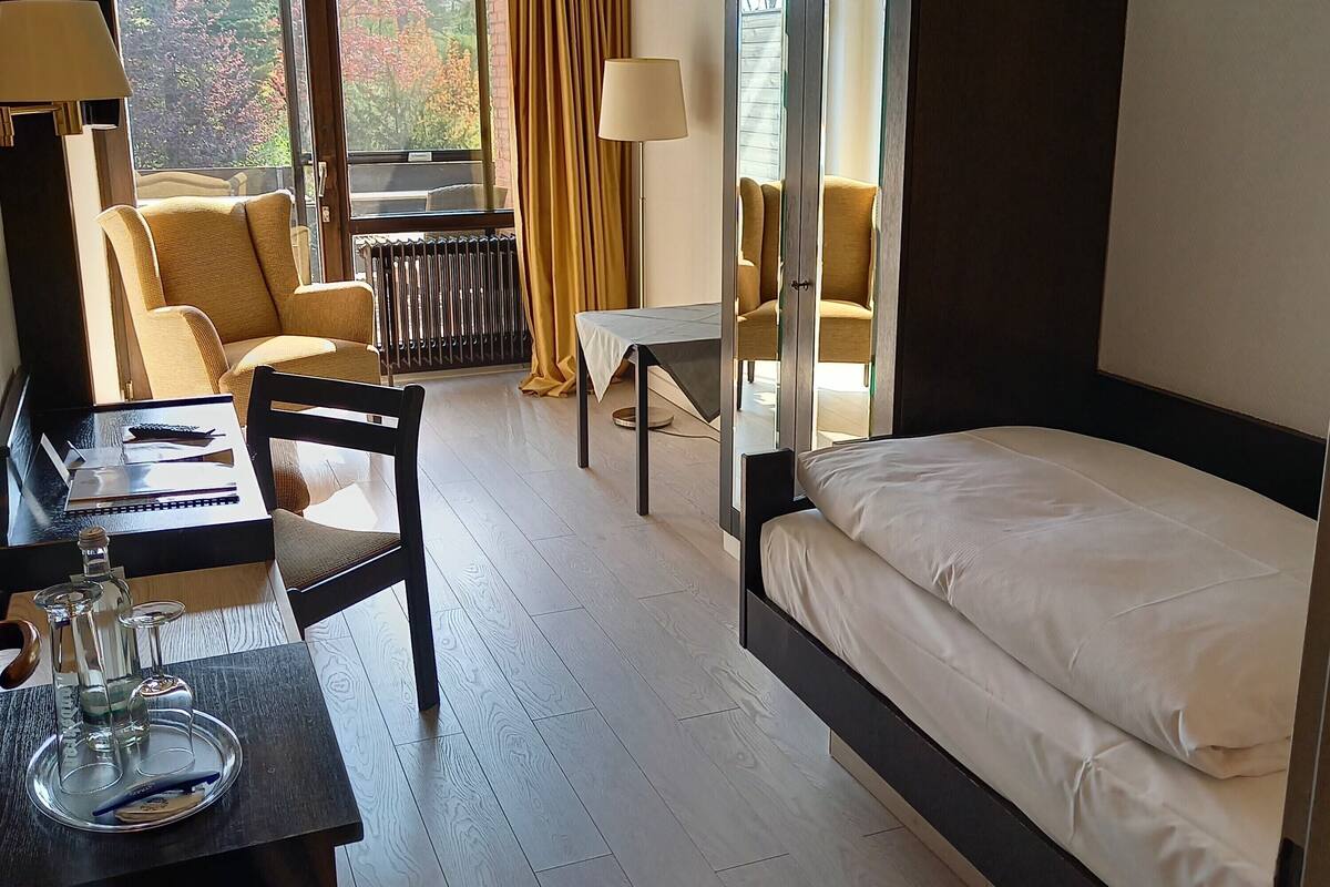 Ein Hotelzimmer mit einem Bett, einem Schreibtisch mit Lampe, einem Sessel, einem Tisch und Blick auf Bäume.