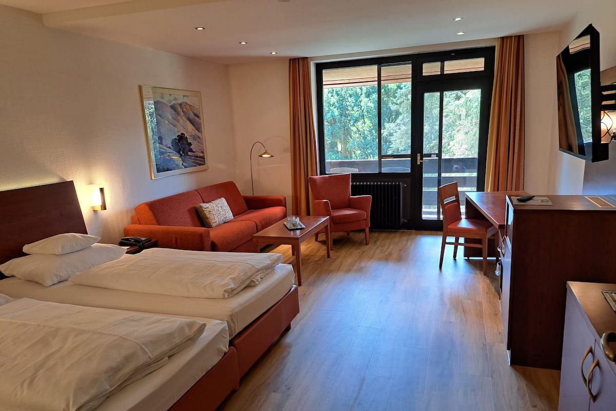 Ein Hotelzimmer mit zwei Betten, einer Schlafcouch, einem Schreibtisch und einem Stuhl. Es gibt ein Fenster mit Vorhängen, ein Bild an der Wand und eine Heizung.