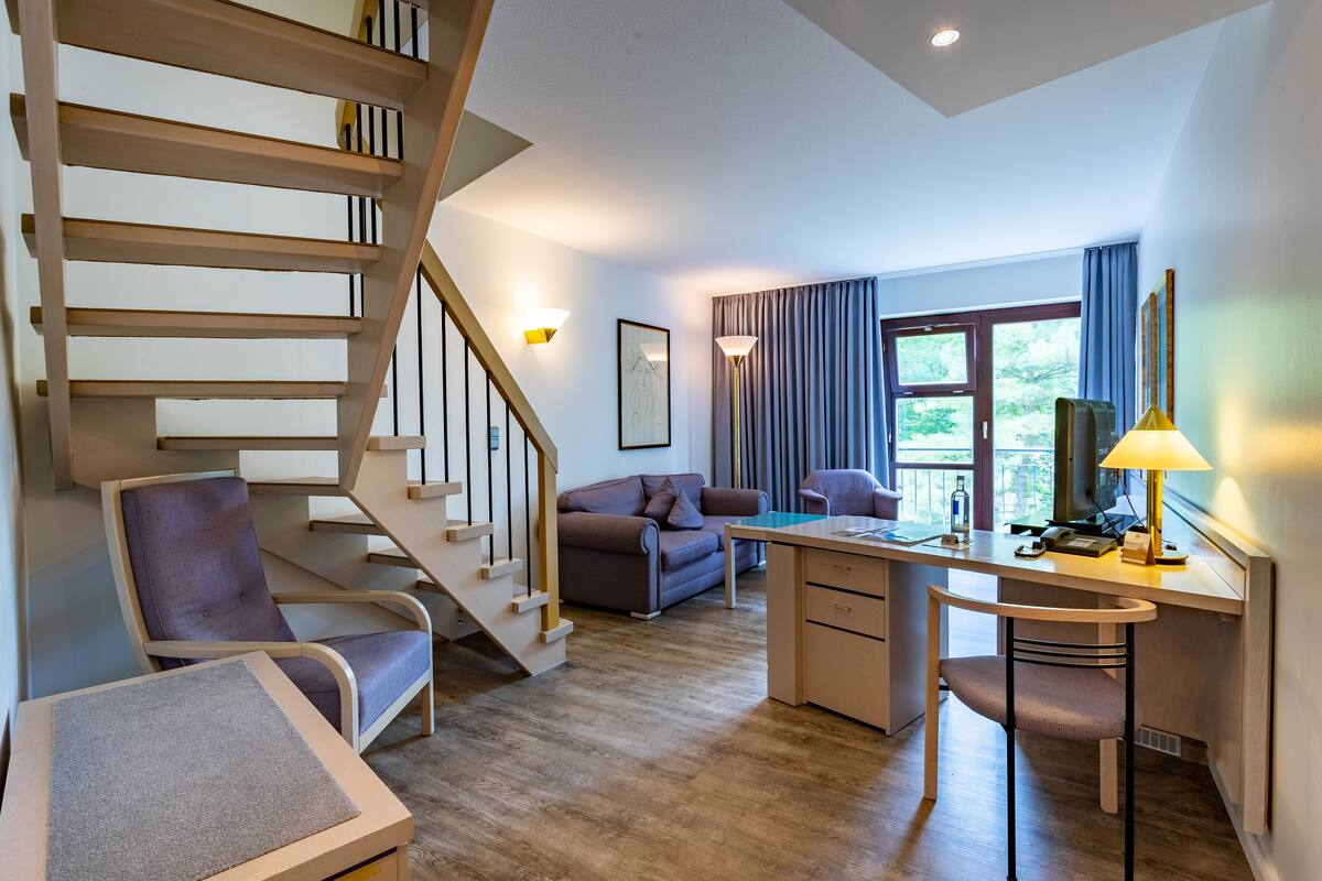 Ein modernes Wohnzimmer mit einer Holztreppe, einem Schreibtisch mit Stuhl, einer Couch und einem großen Fenster mit Vorhängen.