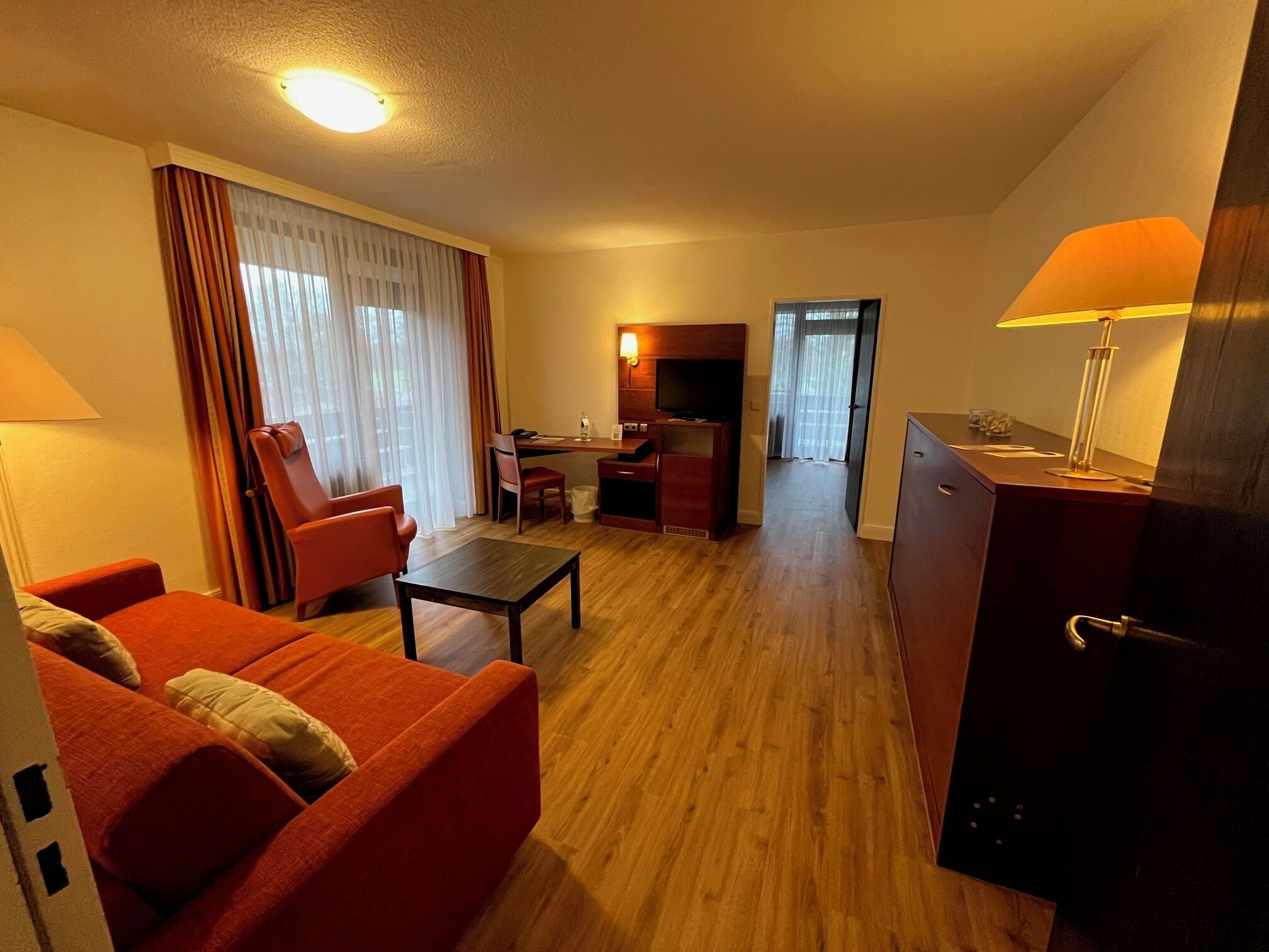 Habitación de hotel con piso de madera, un sofá, un escritorio con silla, televisor, lámpara y armario.