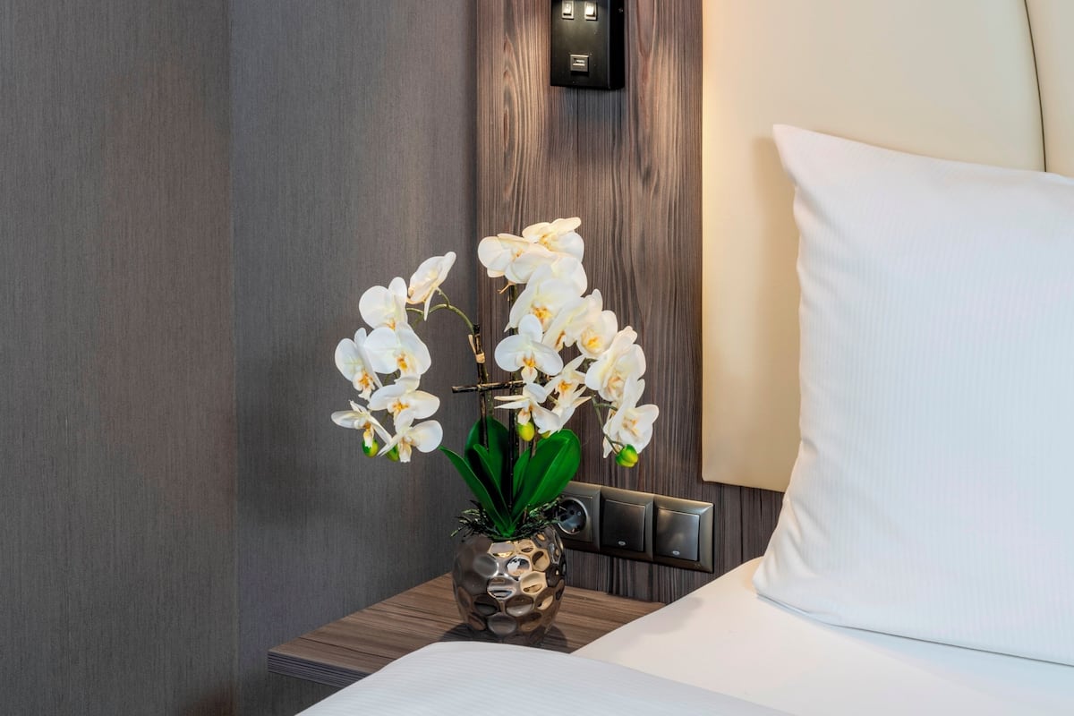 Een hotelkamer met een bed, een nachtkastje met een vaas witte orchideeën en een bedieningspaneel aan de muur.