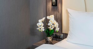 A hotel room with a bed, a nightstand with a vase of white orchids, and a wall-mounted control panel.