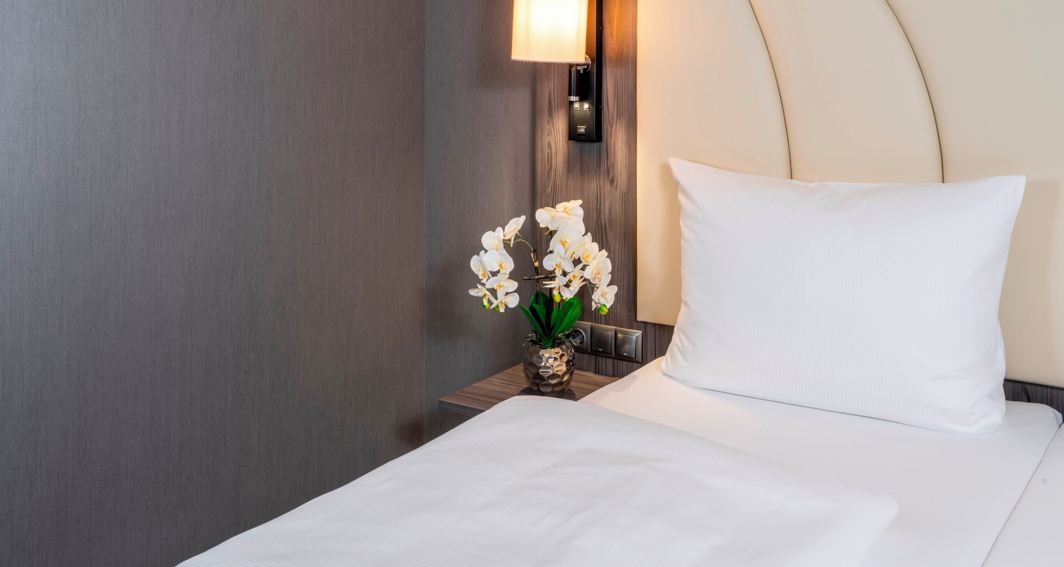 A hotel room with a neatly made bed, a wall-mounted nightstand with a vase of white orchids, and a wall-mounted lamp.