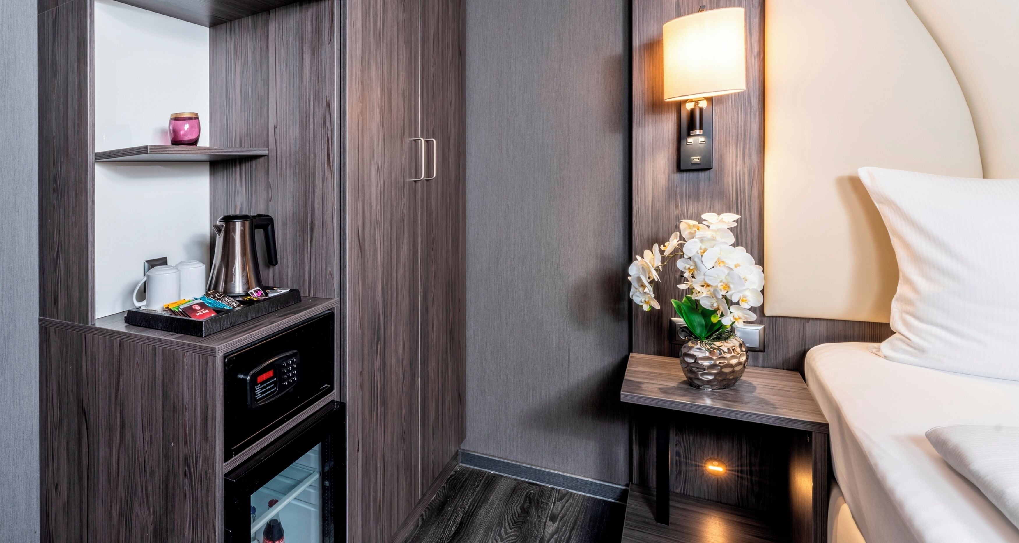 A hotel room with a bed, a nightstand with a lamp, a coffee maker, and a small shelf with a pink candle.