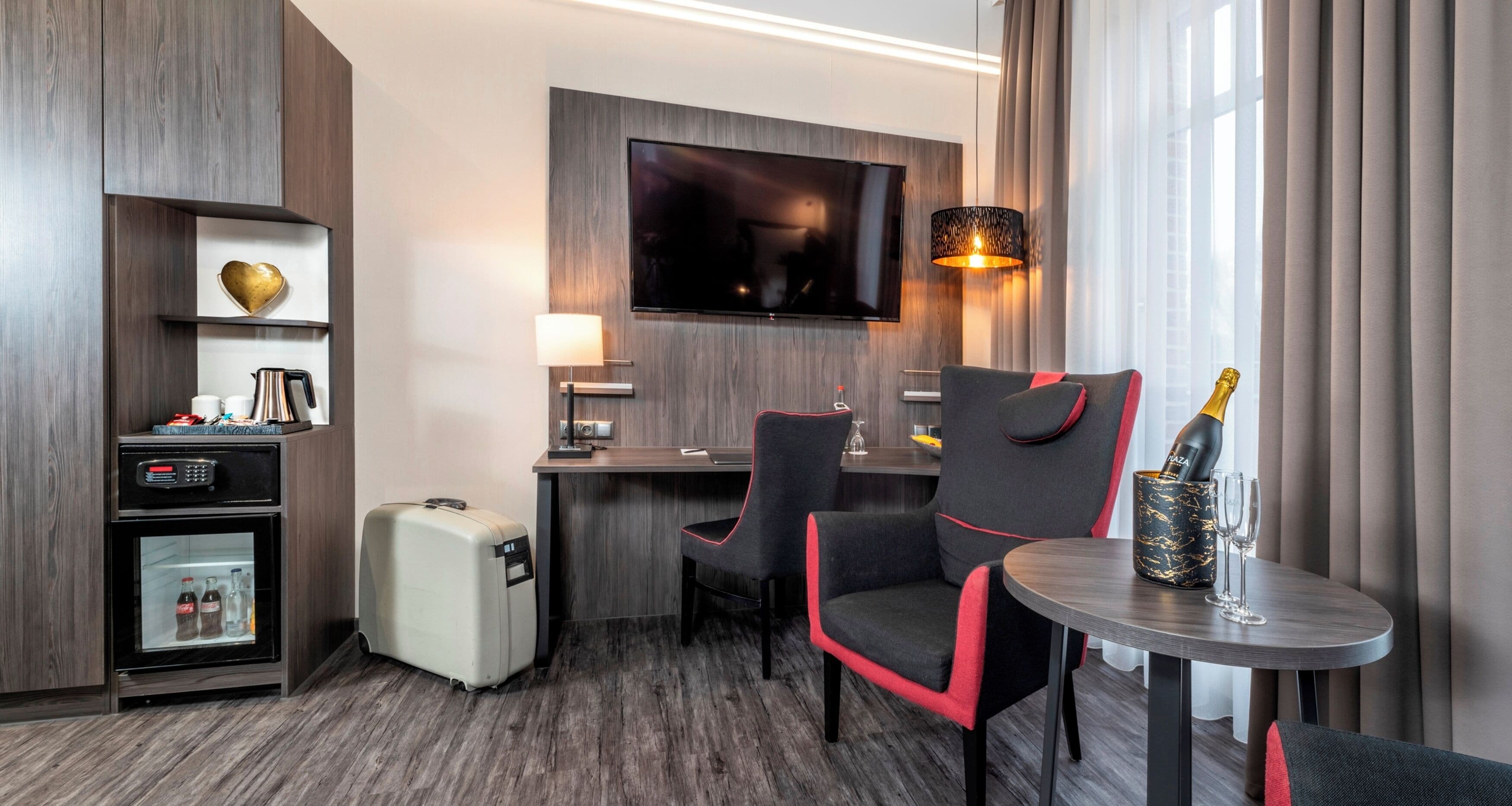 Et hotelværelse med et fladskærms-tv, et skrivebord med lampe, minibar og et siddeområde med et bord, samt en minibar med en champagneflaske.