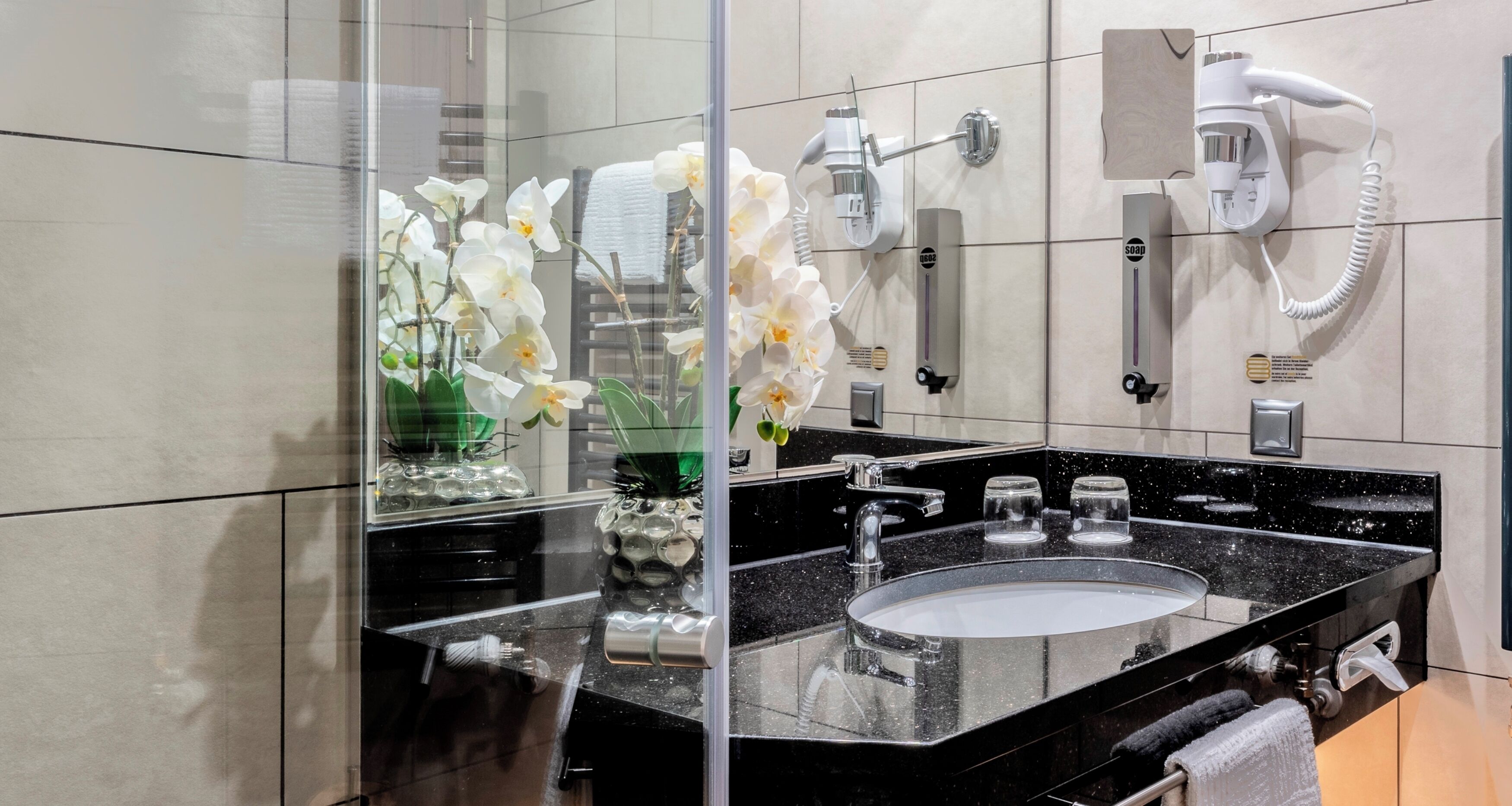 Un baño moderno con un lavabo de granito negro, dos secadores de pelo montados en la pared y una jarra de cristal con orquídeas blancas.