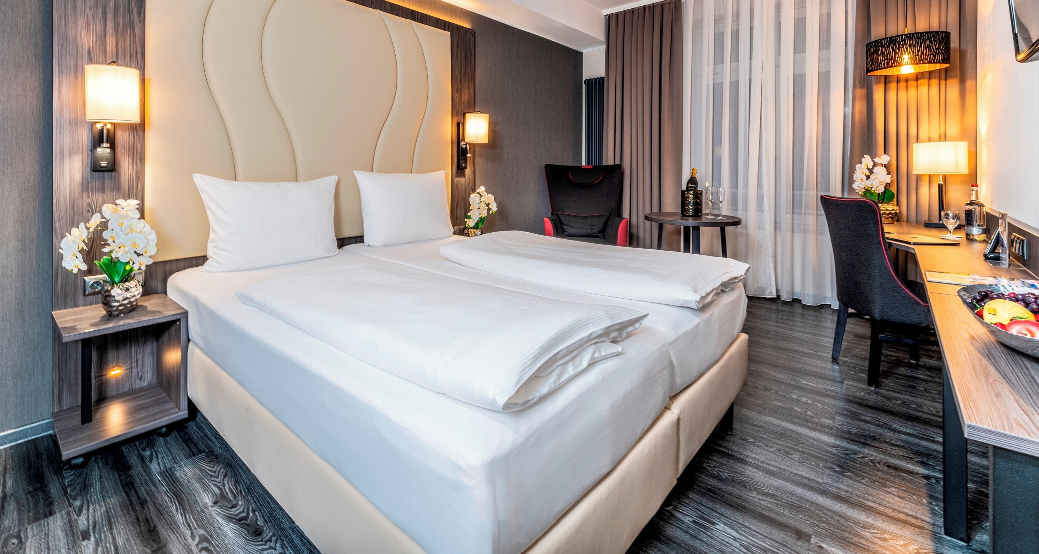 Una habitación de hotel con cama doble, dos mesitas de noche con lámparas, un escritorio pequeño con una silla y vistas a la ciudad a través de amplios ventanales.