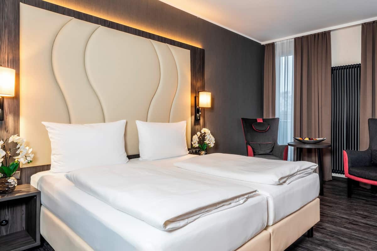 Hotelkamer met een groot bed, twee nachtkastjes, een stoel, een klein tafeltje met een fruitschaal, en een raam met gordijnen.