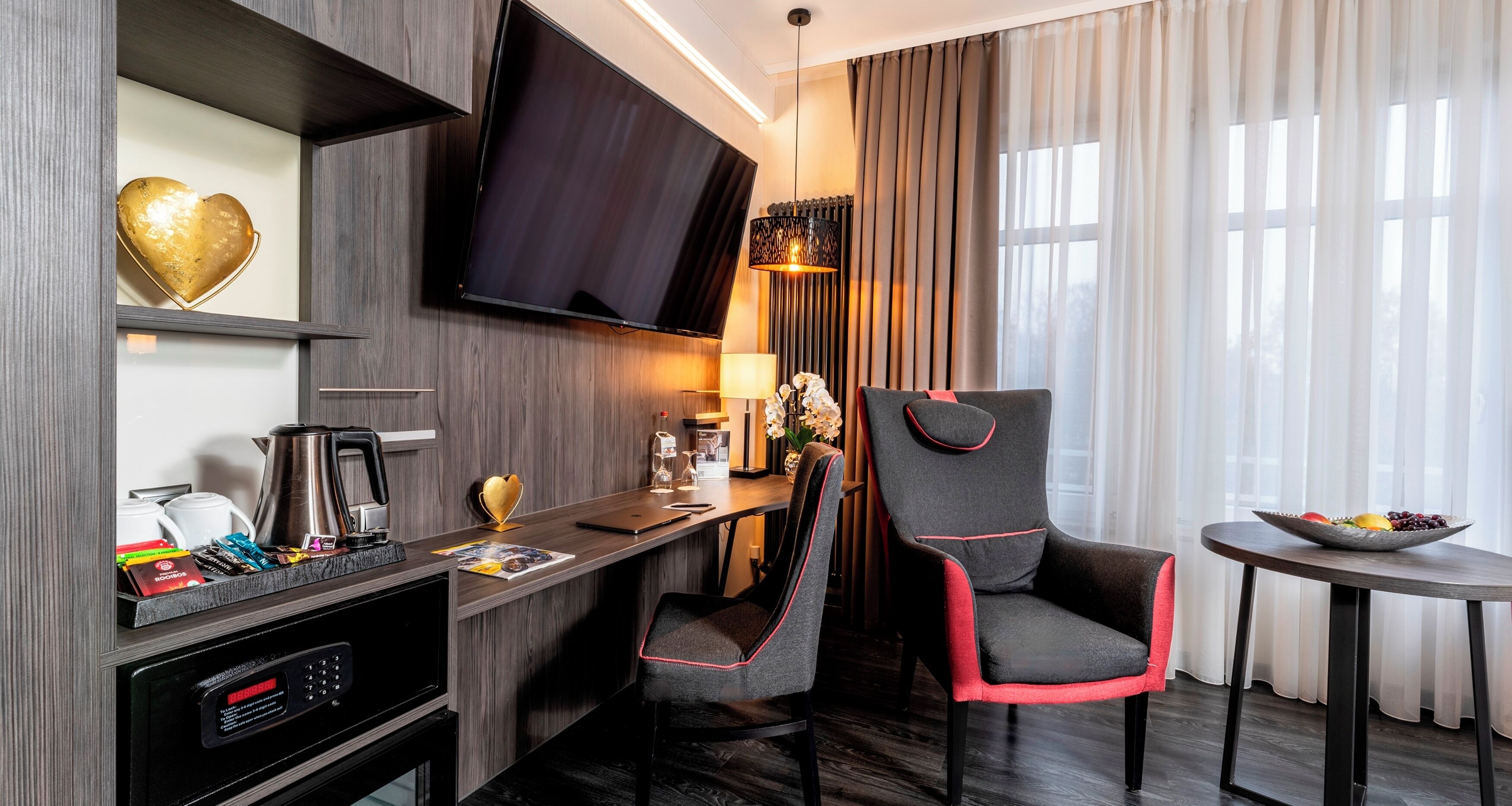 A modern hotel room with a flat-screen TV, a desk with a coffee maker, a chair, and a small table with fruit.