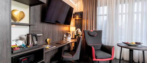 A modern hotel room with a flat-screen TV, a desk with a coffee maker, a chair, and a small table with fruit.