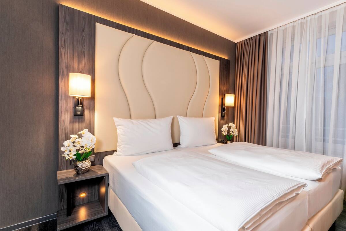Een hotelkamer met een groot bed, twee nachtkastjes, een wandlamp en uitzicht op het landschap door dunne gordijnen.