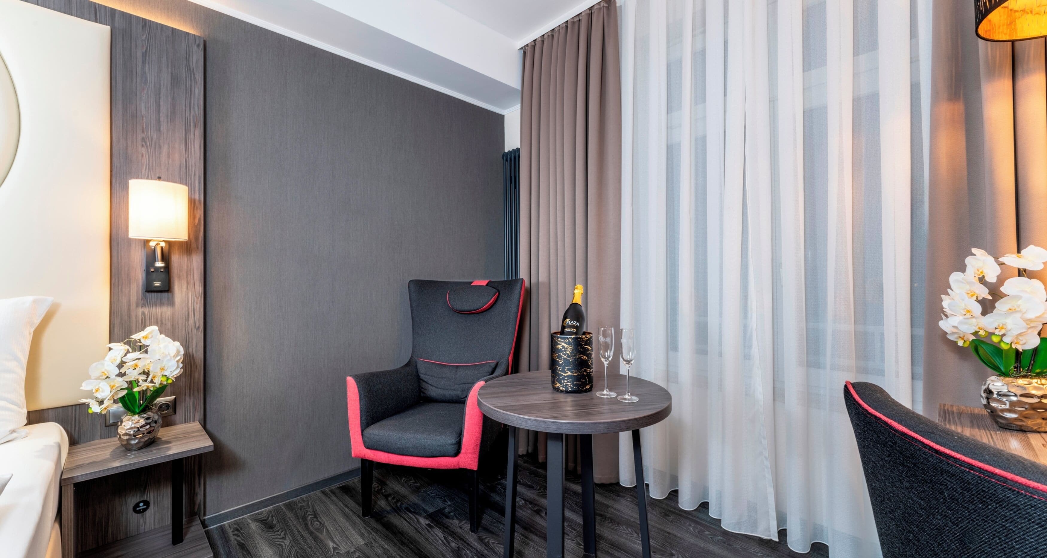 Habitación de hotel con una butaca tapizada de gris, una mesita redonda con una botella y vasos, un florero con flores blancas y una lámpara sobre una mesita de noche.