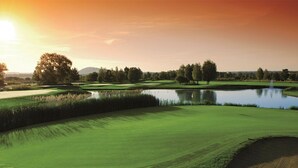 Un campo de golf con un estanque, árboles y un atardecer.
