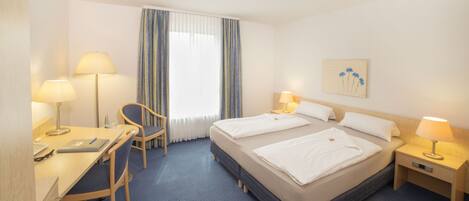A hotel room with two beds, a desk with a chair, a lamp, and a window with curtains.