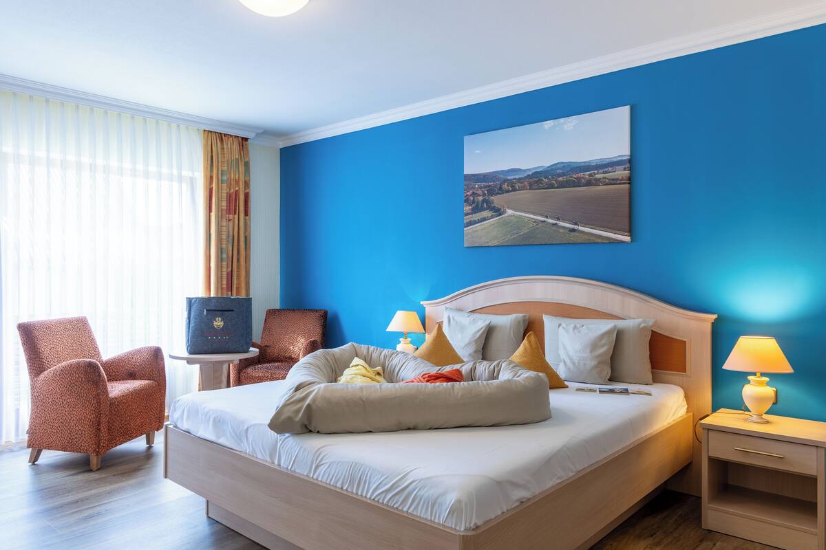 Habitación de hotel con una cama grande, dos sillas, una mesita de noche con lámpara, una pequeña mesa y un cuadro en la pared.