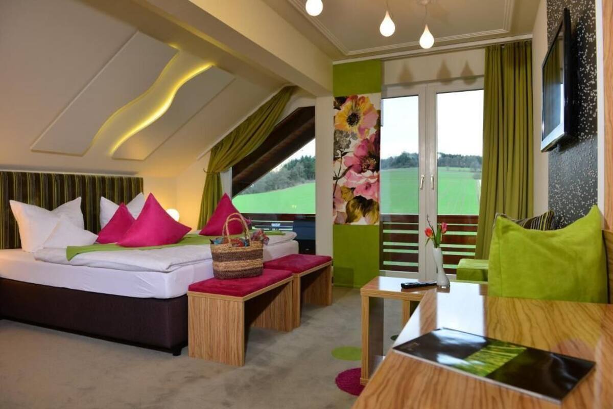 Un dormitorio con cama, mesitas de noche, una pequeña mesa y vista a un paisaje verde por la ventana.