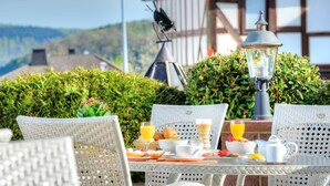 A breakfast table set outdoors with wicker chairs, a lamp, and a view of a mountain and buildings in the background. - Hotel Freund (Voehl)