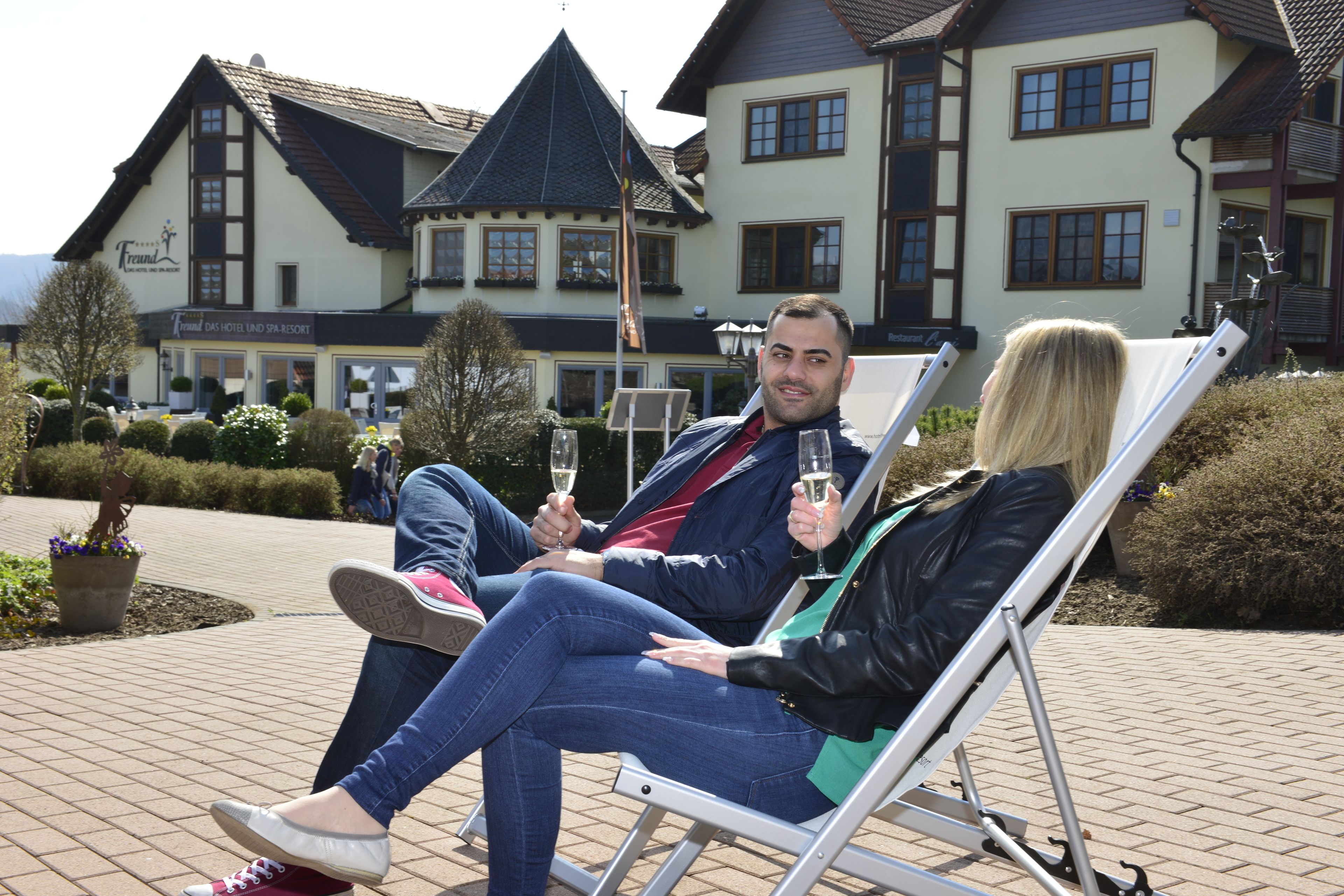 A couple relaxing on a sun lounger in front of a hotel with a sign reading "Hotel Das Hotel und Spa Resort".