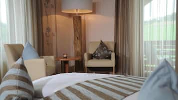 Quarto de hotel com cama, duas cadeiras, uma mesa, luminária e vista para um campo verde pela janela.