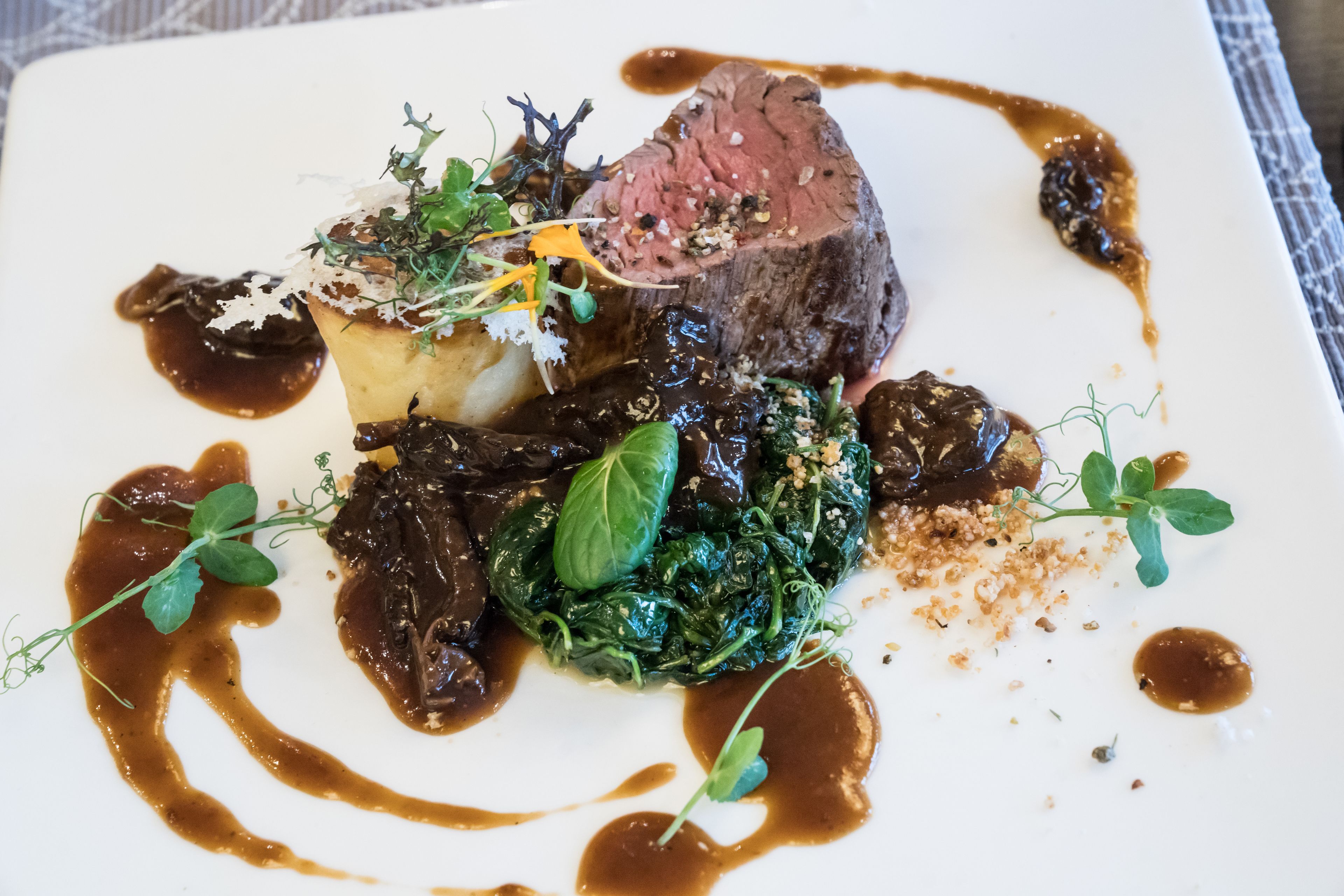 Un plato cuadrado con un trozo de carne, una patata al horno y vegetales verdes, adornado con hierbas y salsa.
