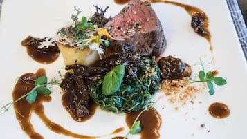 Un plato cuadrado con un trozo de carne, una patata al horno y vegetales verdes, adornado con hierbas y salsa.