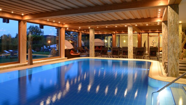 An indoor swimming pool with a wooden ceiling and large windows overlooking a garden. - Hotel Freund (Voehl)