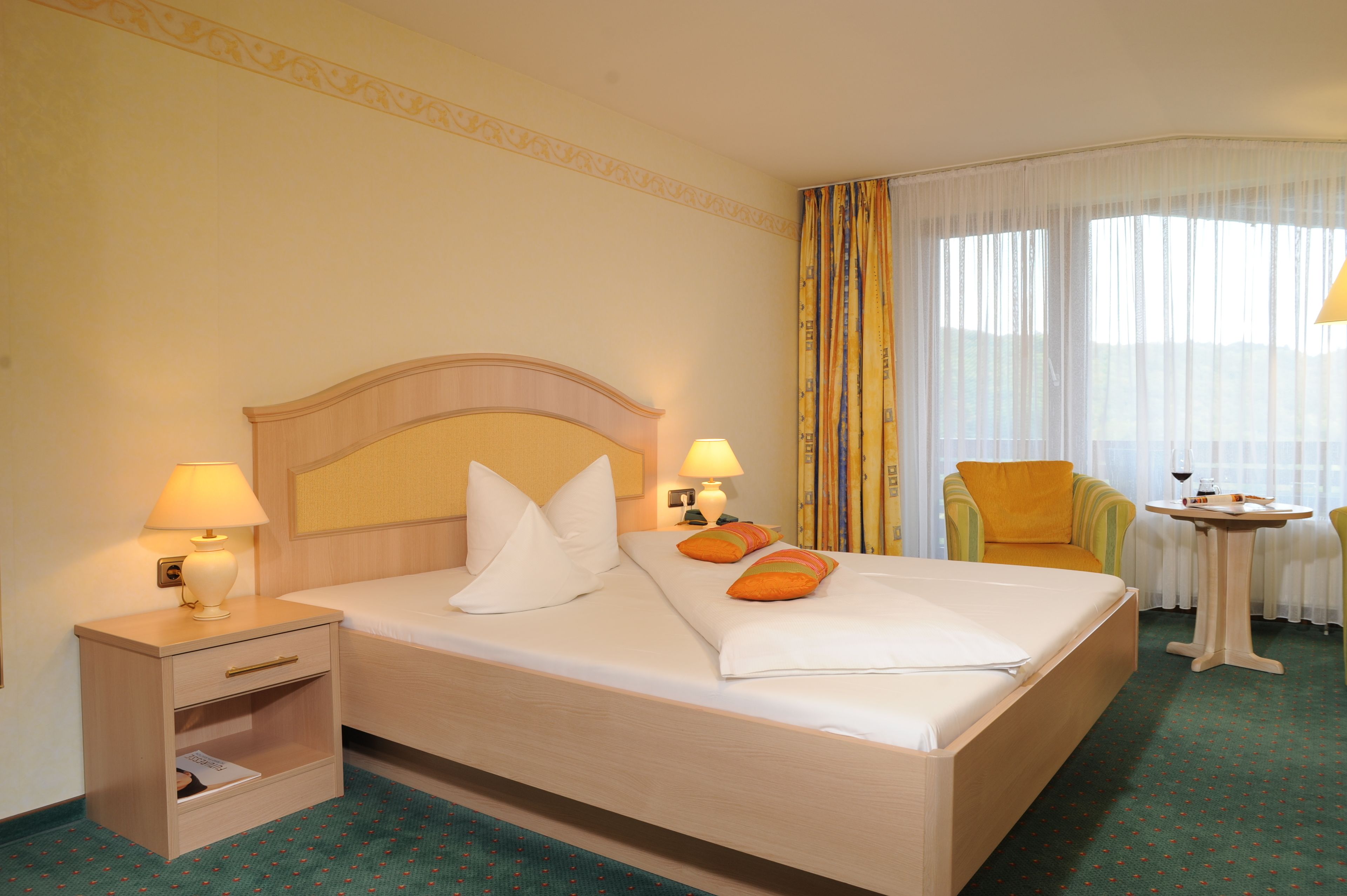 Quarto de hotel com cama, duas luminárias de cabeceira, uma cadeira, uma mesa pequena com uma taça de vinho e uma janela com cortinas.