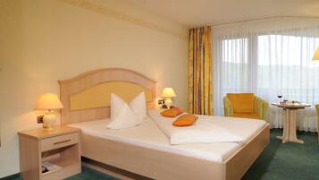 Quarto de hotel com cama, duas luminárias de cabeceira, uma cadeira, uma mesa pequena com uma taça de vinho e uma janela com cortinas.