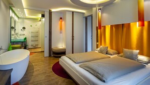 A modern hotel room with a large bed, a separate seating area, a bathtub, and a well-lit ceiling. - Hotel Freund (Voehl)