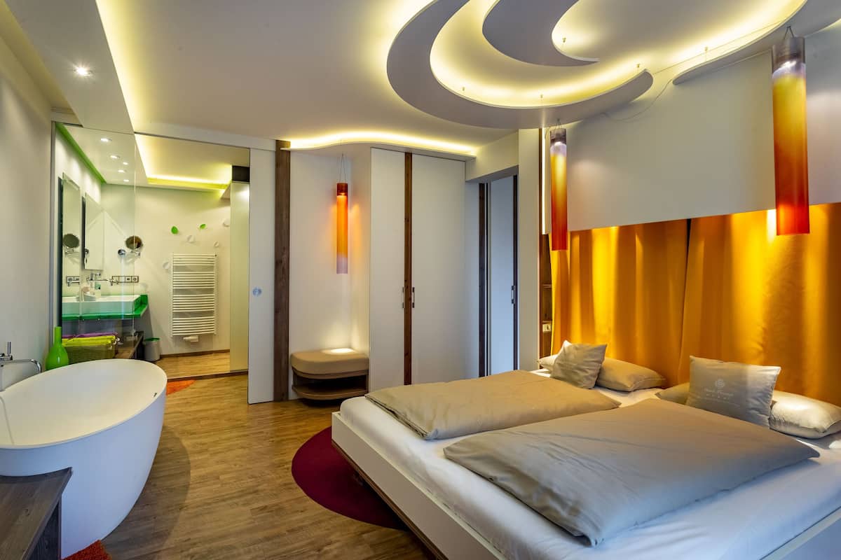 Una habitación de hotel moderna con una cama grande, una zona de estar separada, una bañera y un techo bien iluminado.