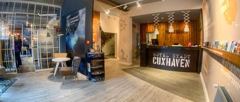 A hotel lobby with a reception desk labeled "HOTEL STAADT CUXHAVEN," a wooden wall with a star decoration, and a seating area with a round table and chairs.