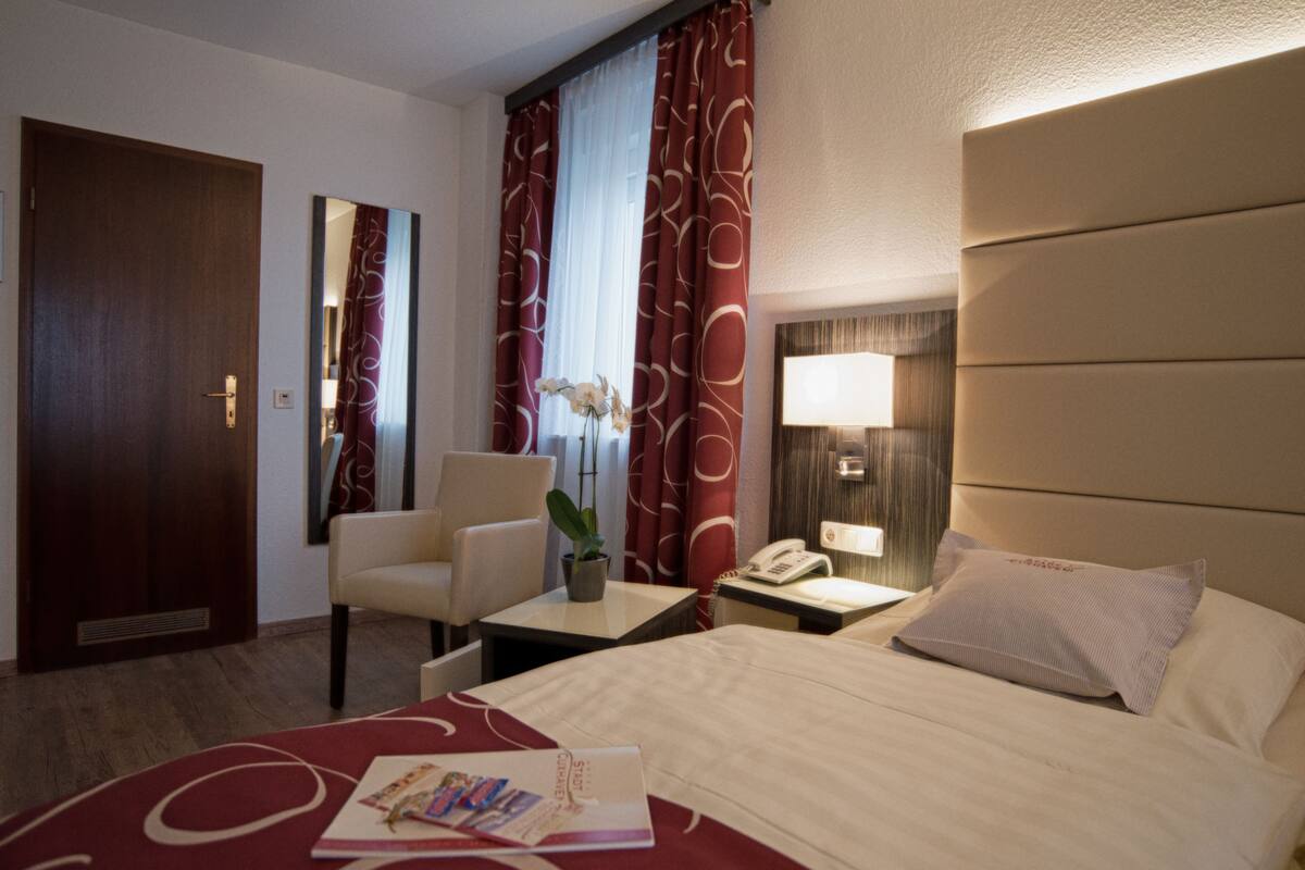 A hotel room with a bed, a desk, a chair, a nightstand with a lamp, a telephone, and a mirror.