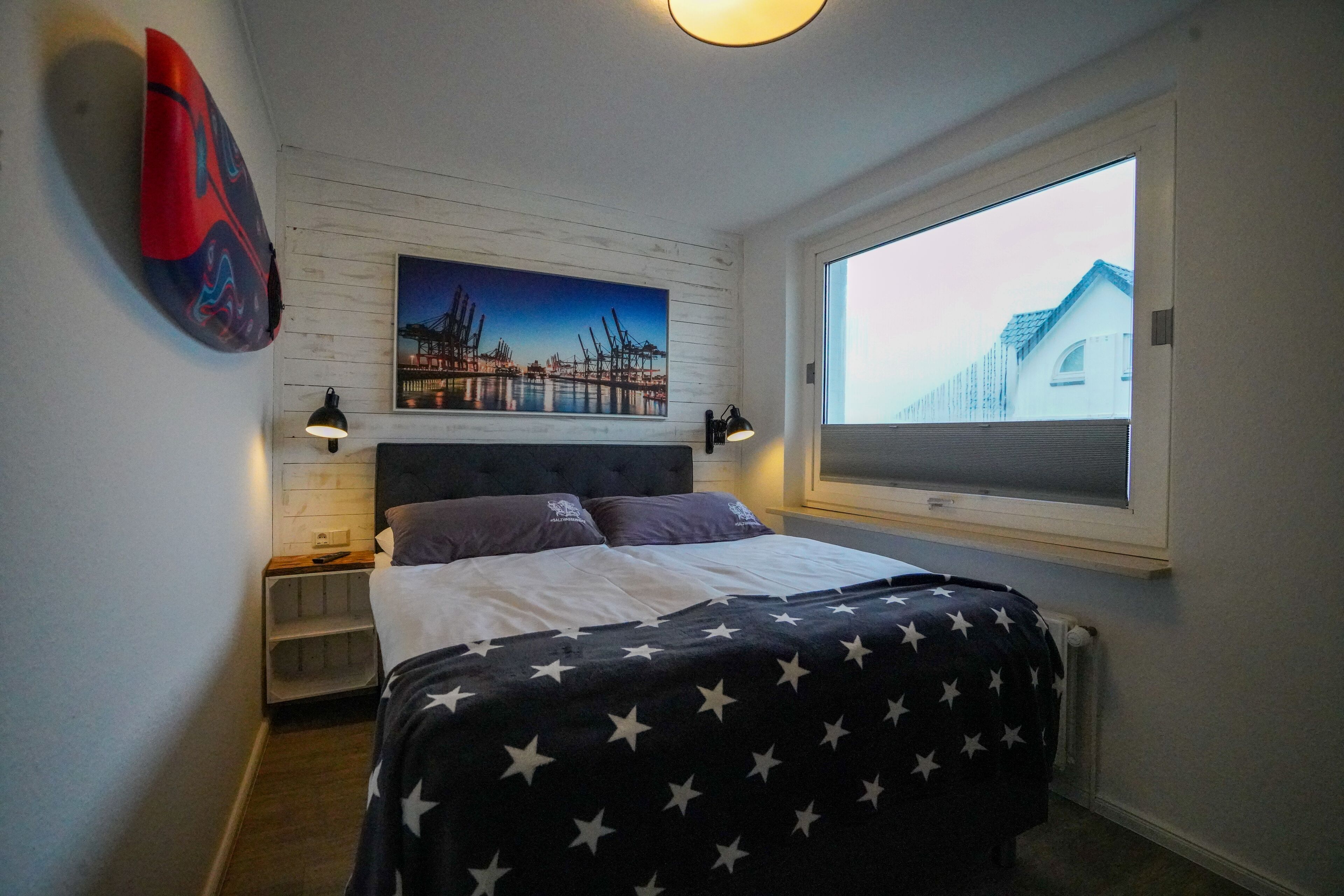 Une chambre à coucher avec un lit, une table de chevet, une lampe fixée au mur, une fenêtre donnant sur un immeuble et un tableau accroché au mur.