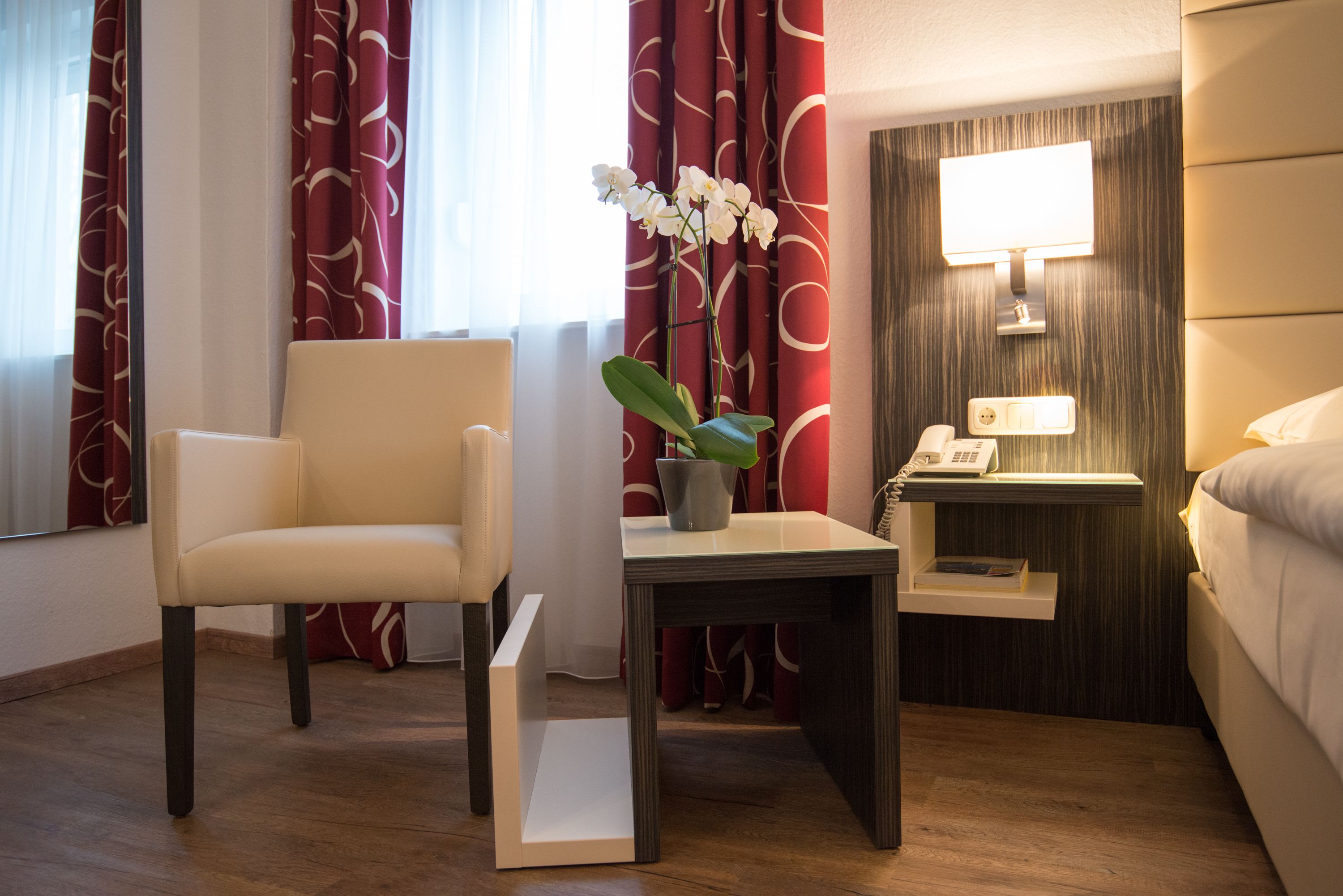 A hotel room with a bed, a bedside table with a lamp, a chair, and a small table with a potted plant.