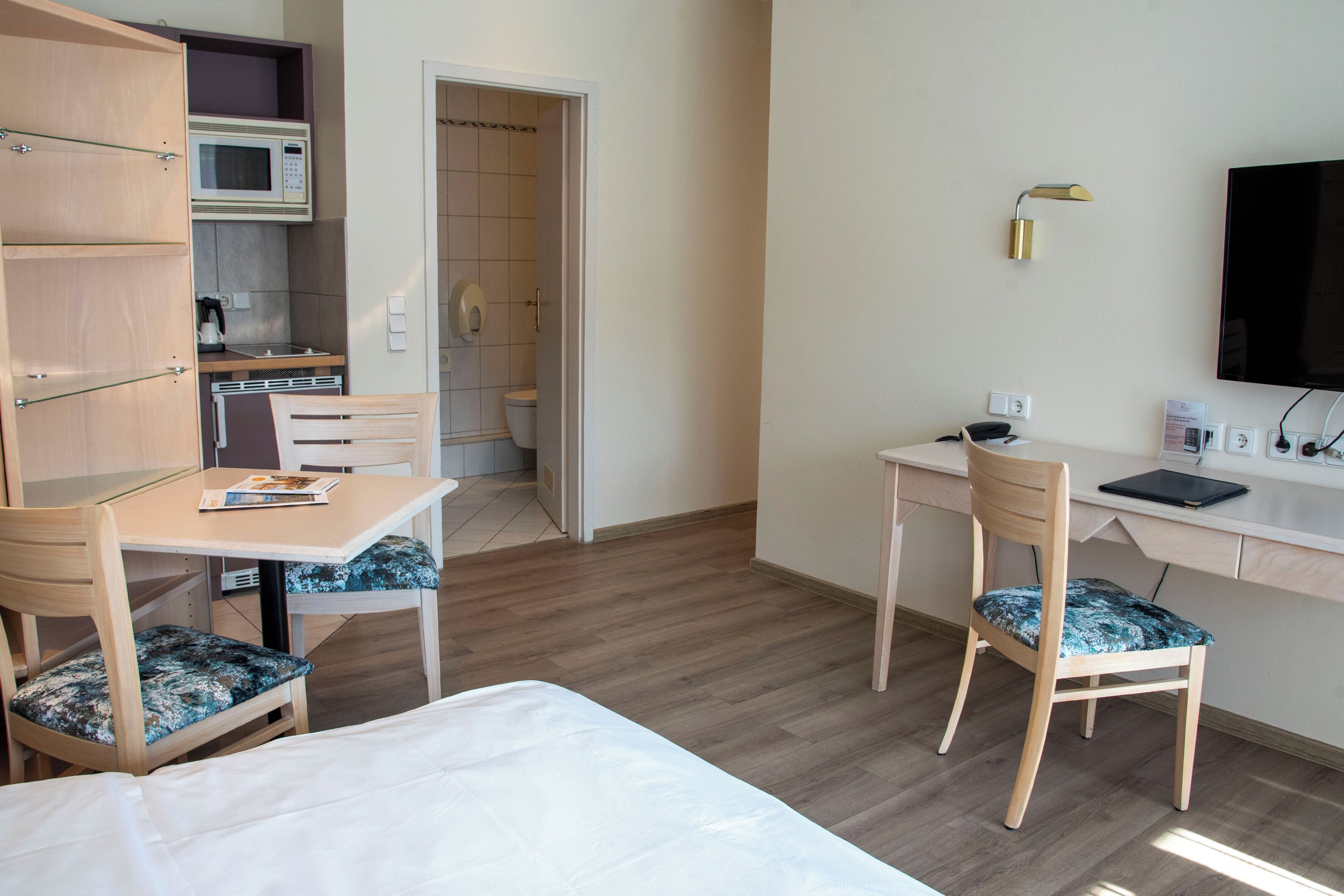 Una habitación de hotel con cama, escritorio con silla, microondas, televisión y baño visible a través de una puerta abierta.