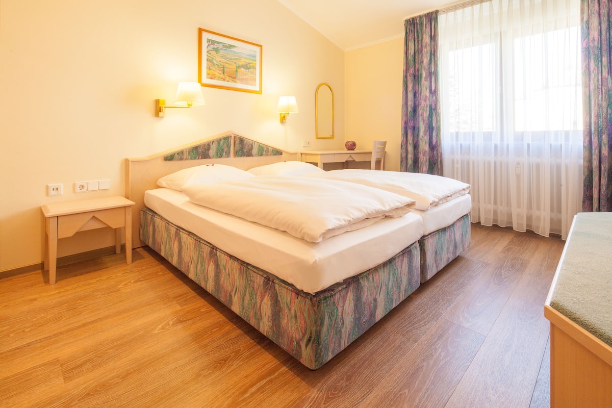 A hotel room with a bed, bedside table, lamp, desk, chair, and a window with curtains.