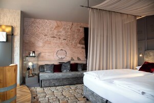 A modern hotel room with a large map on the wall, a grey sofa, a wooden side table, and a neatly made bed.