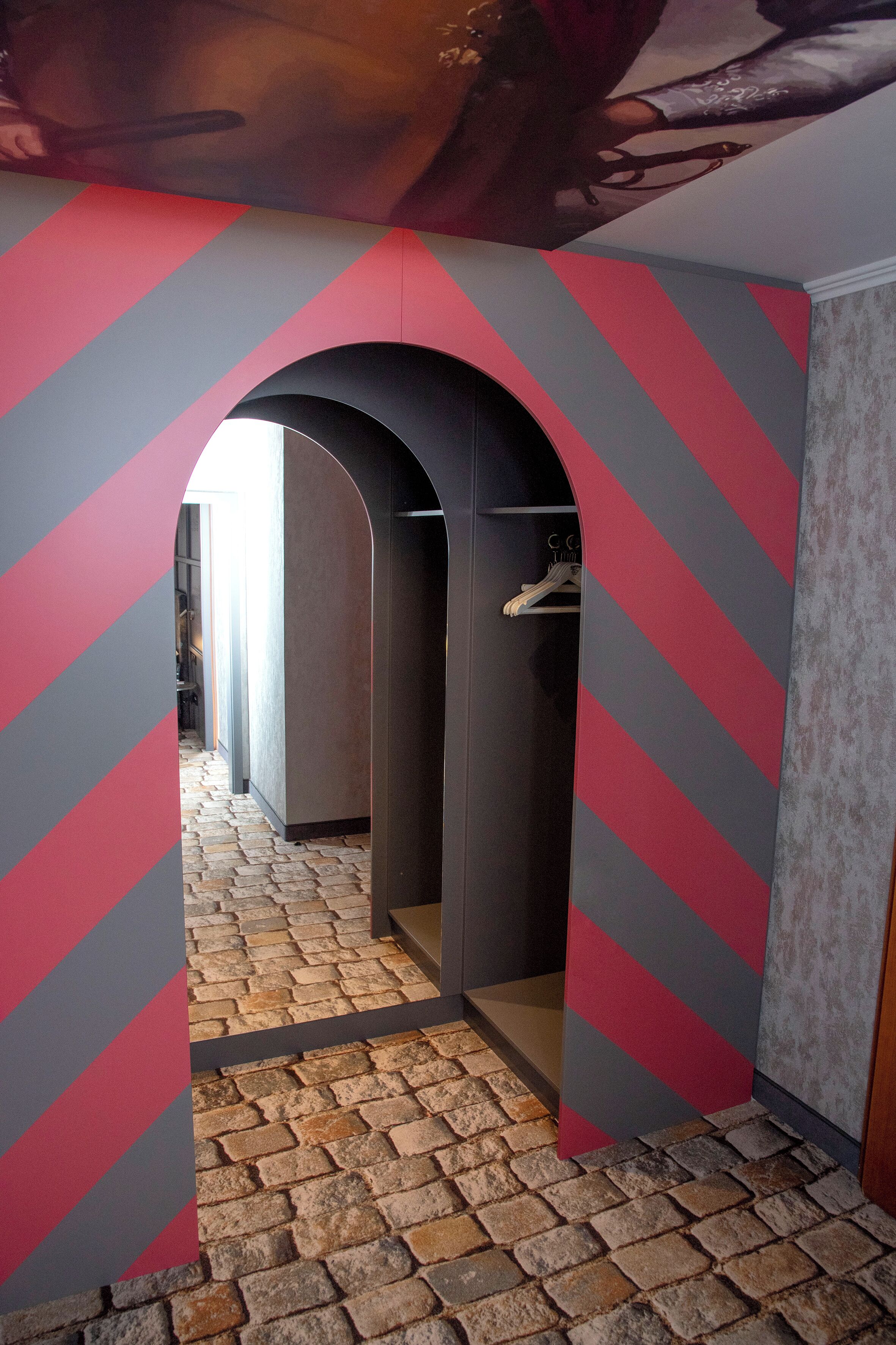 Ein Korridor mit Terrazzoboden, einer Wand in Rot und Grau gestreift und einem schwarzen Bogen, der in einen Raum mit einer Kleiderstange führt.
