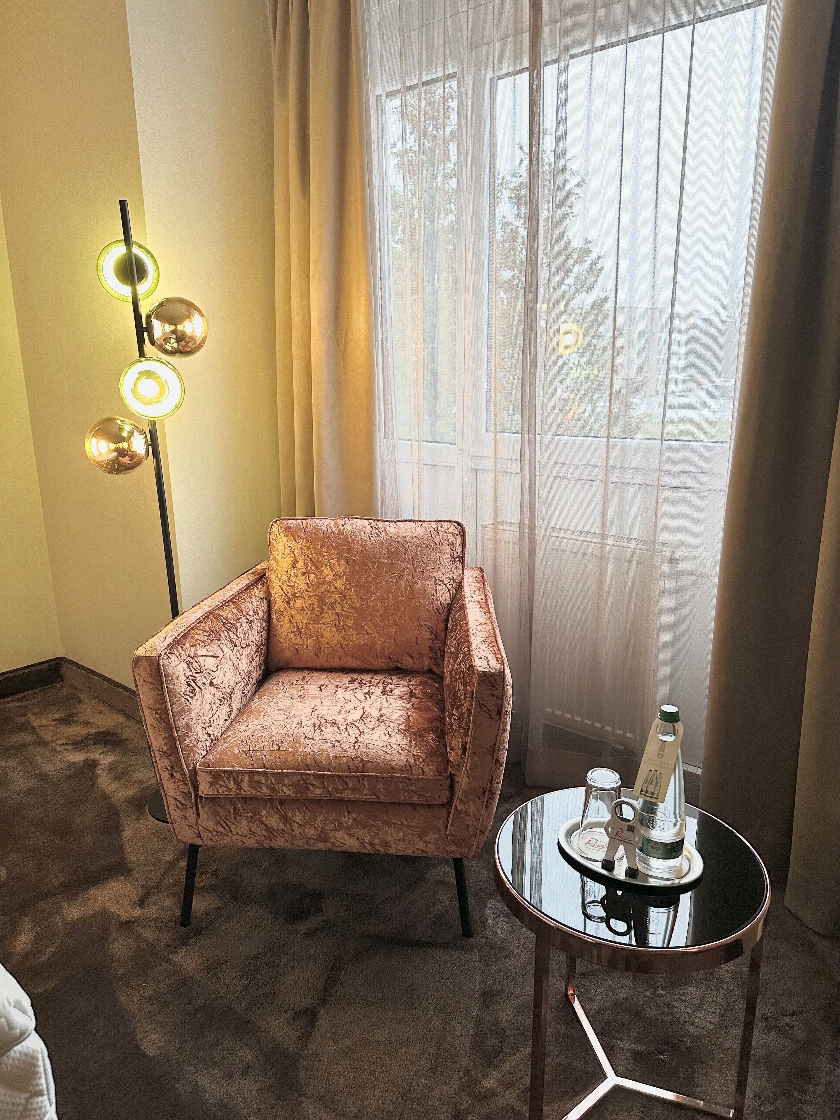 A room with a cushioned armchair, a small round table with a bottle and glass, and a floor lamp with three spherical lights.