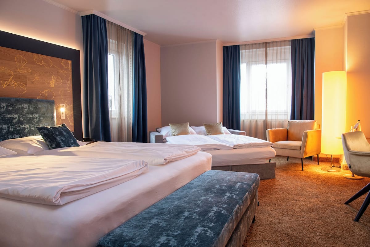A hotel room with two beds, a sofa, a chair, a lamp, and a large window with curtains.