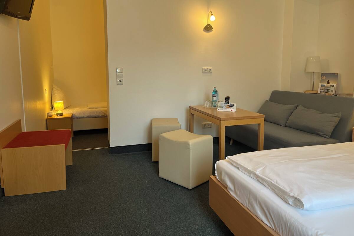 A hotel room with a bed, a small table with a remote control, a sofa, a nightstand with a lamp, and a wall-mounted TV.
