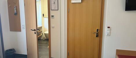 A hotel room with a wooden door, a small desk, a chair, a trash bin, and a wall-mounted TV.