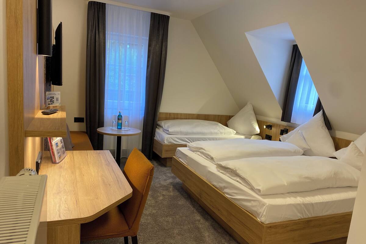 A hotel room with a desk, two beds, a small round table with a bottle, and a TV mounted on the wall.