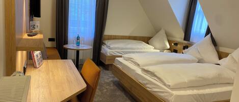 A hotel room with a desk, two beds, a small round table with a bottle, and a TV mounted on the wall.
