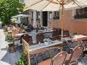 Un café al aire libre con mesas y sillas de madera, sombrillas y plantas en macetas.