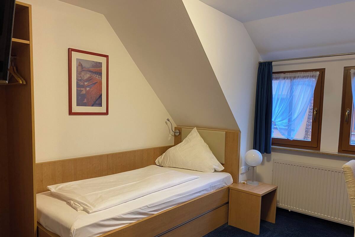 A single bed with a wooden headboard, a bedside table with a lamp, a framed picture on the wall, and a window with curtains.