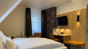 A hotel room with a bed, a desk, a chair, a TV mounted on the wall, and a window with curtains. - AKZENT Hotel Schranne (Rothenburg ob der Tauber)