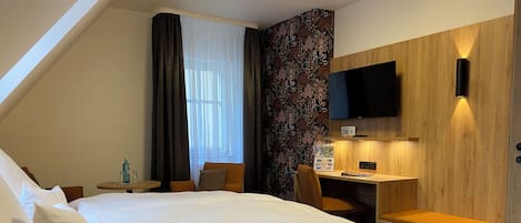 A hotel room with a bed, a desk, a chair, a TV mounted on the wall, and a window with curtains.