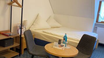 A compact hotel room with a bed, a small round table with a remote control, a chair, a desk with a chair, and a wardrobe with a hanger.